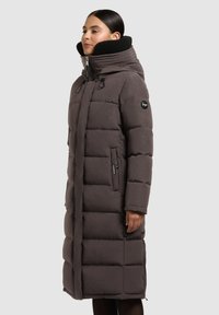 Cappotto imbottito lungo di un grigio scuro con colletto alto, caratterizzato da fodera in pelliccia sintetica nera, dettagli con zip e tasche, e una vestibilità comoda.