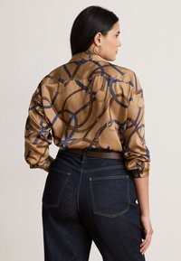 Γυναίκα με σκούρα μαλλιά που φοράει χρυσά σκουλαρίκια κρίκους, ένα καφέ εμπριμέ μακρυμάνικο πουκάμισο και σκούρο τζιν με ψηλή μέση, με φορά πλάγια.