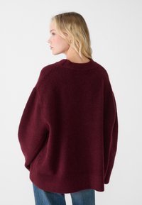 Pull oversized bordeaux avec une finition douce et texturée, un col côtelé et des épaules tombantes. Associé à un jean bleu clair.