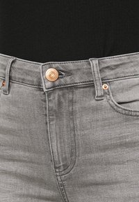 Acercamiento a la cintura de unos pantalones vaqueros de mezclilla gris con botón de cobre y detalles de costura, combinados con una blusa negra.