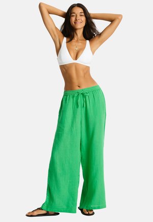 Kobieta w białym krótkim topie i luźnych zielonych spodniach, stojąca z uniesionymi rękami i uśmiechająca się na jednolitym tle.