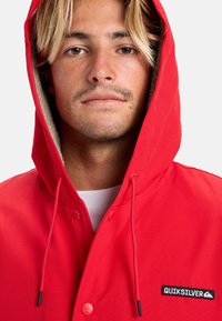 Rød hættejakke med fleece-foret indvendigt, med sorte knapper og et Quiksilver-logo patch på brystet.
