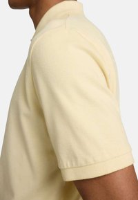 Polo shirt jaune pâle avec une finition texturée, à manches courtes et col classique. Possède une coupe nette et ajustée au niveau de l'épaule.
