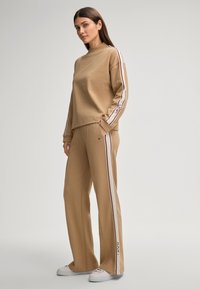 Beige Sweatshirt- und Hosen-Set mit kontrastierenden Seitenstreifen, aus weichtexturiertem Stoff und mit lässigem Schnitt. Weiße Sneaker runden das Outfit ab.