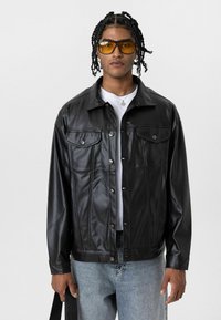 Zwarte jas van nepleer met een kraag, twee borstzakken, knopen aan de voorkant en een gladde textuur, gecombineerd met blauwe spijkerbroek.