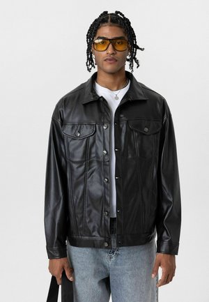 Zwarte jas van nepleer met een kraag, twee borstzakken, knopen aan de voorkant en een gladde textuur, gecombineerd met blauwe spijkerbroek.