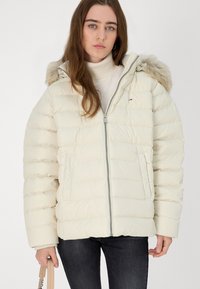 Veste matelassée crème avec capuche en fourrure amovible, fermeture éclair et motif matelassé horizontal. Poches latérales et texture douce.