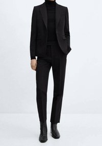 Person in einem taillierten schwarzen Blazer, schwarzem Rollkragenpullover, maßgeschneiderten schwarzen Hosen und schwarzen Ankle-Boots, steht vor einem schlichten hellen Hintergrund.