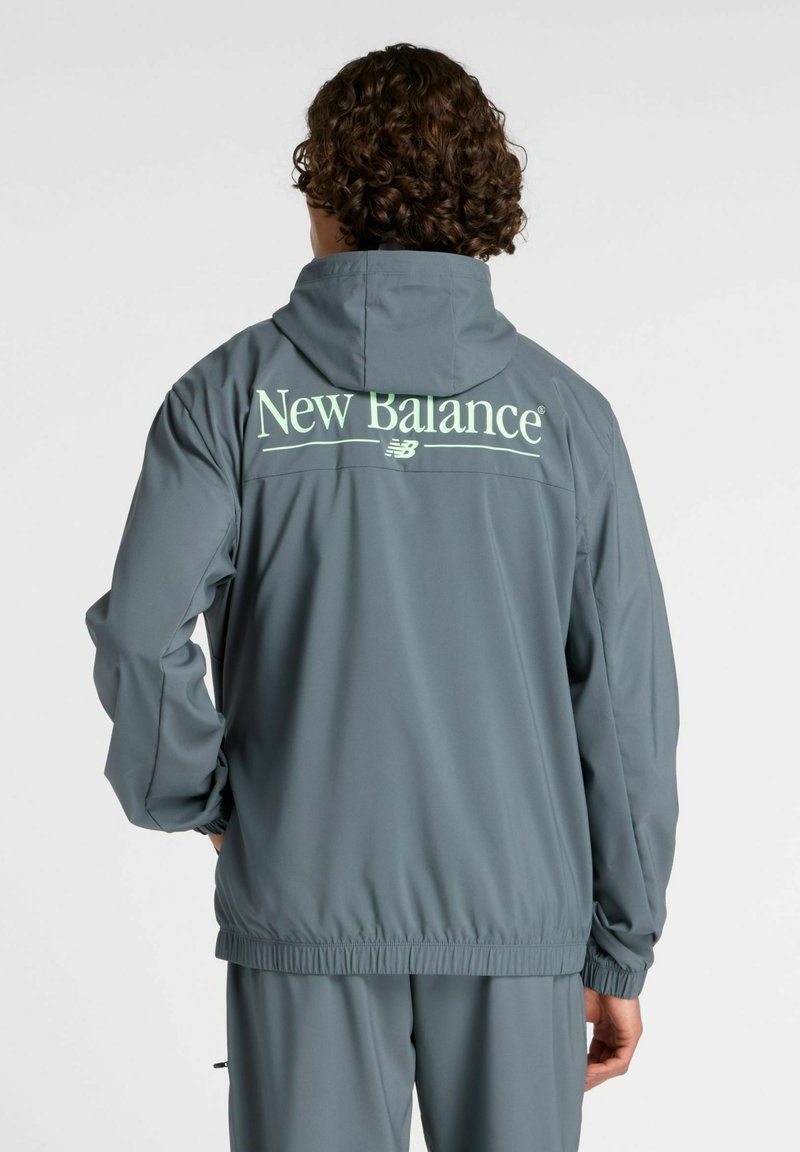 Man met krullend haar die een grijze New Balance hoodie en bijpassende broek draagt, met zijn gezicht naar weg gericht tegen een effen witte achtergrond.