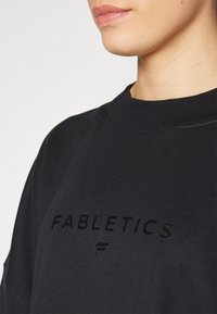 Sudadera negra de Fabletics con cuello redondo y estampado de logo texturizado. La tela parece suave, con un corte casual y elementos de diseño mínimos.