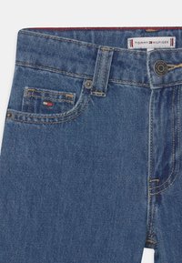 Jasnoniebieskie dżinsy z klasycznym pięciopocketowym wzorem, widocznymi szwami i małą metką z logo. Tekstura jest gładka, z lekkim przetarciem.