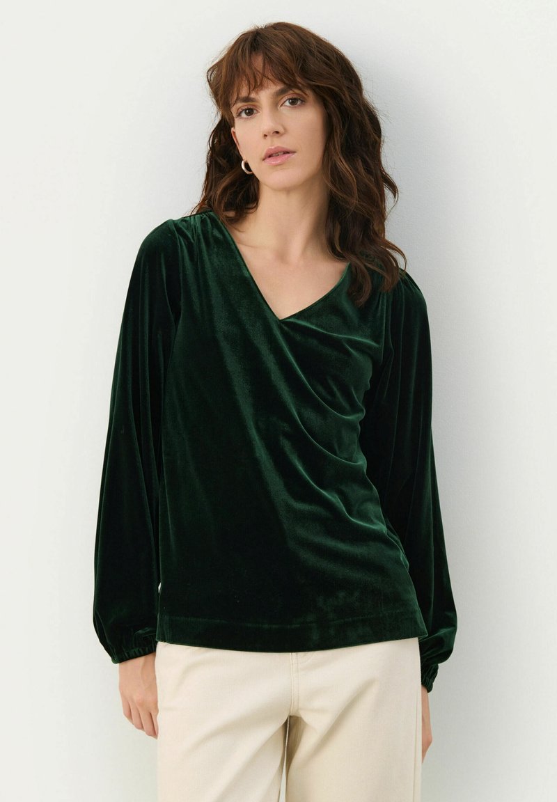 Grüne Samtbluse mit V-Ausschnitt. Sie verfügt über lange, ballonförmige Ärmel und ein drapiertes Vorderdesign. Glatte Textur und reiche Farbe.