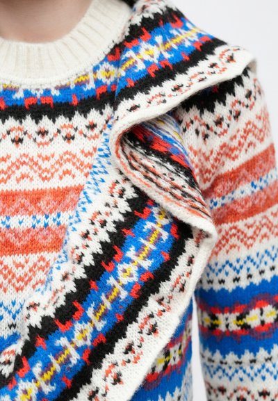 Χρωματιστό πλεκτό πουλόβερ με συνδυασμό κόκκινων, μπλε, λευκών και μαύρων σχεδίων· ριμπ λαιμόκοψη· λεπτομέρεια διπλωμένου γιακά ορατή.