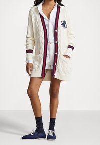 Stickad cardigan i krämfärg med ett kabelmönster och röd och marinblå rand. Har ett lejonemblem, fickor och knappar. I kombination med vit skjorta och marinblå sneakers.