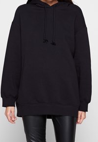 Donna che indossa un felpa nera oversized con lacci abbinata a pantaloni in pelle nera, in piedi davanti a uno sfondo bianco semplice.