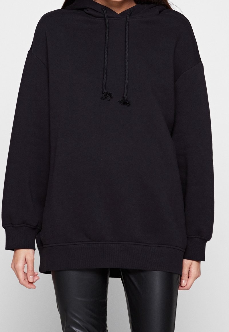 Donna che indossa un felpa nera oversized con lacci abbinata a pantaloni in pelle nera, in piedi davanti a uno sfondo bianco semplice.
