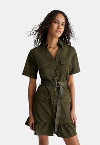 Abito verde oliva con bottoni, colletto, due tasche sul petto, orlo arricciato e una cintura con fibbia circolare. Tessuto leggero.