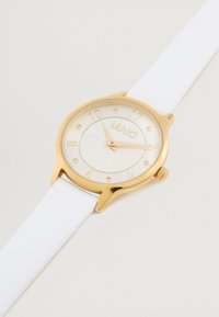 Ceas de mână cu nuanță de aur, curea din piele albă, cadran rotund, dial clar, marcaje de oră aurii și o coronă pe partea dreaptă.