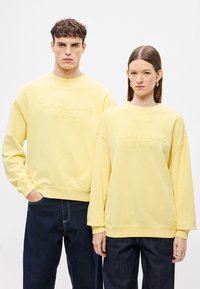 Κίτρινα φούτερ με ανάγλυφο λογότυπο "TOMMY JEANS", με ριμπ μανσέτες και τελείωμα, φορεμένα με σκούρο τζιν από δύο άτομα.