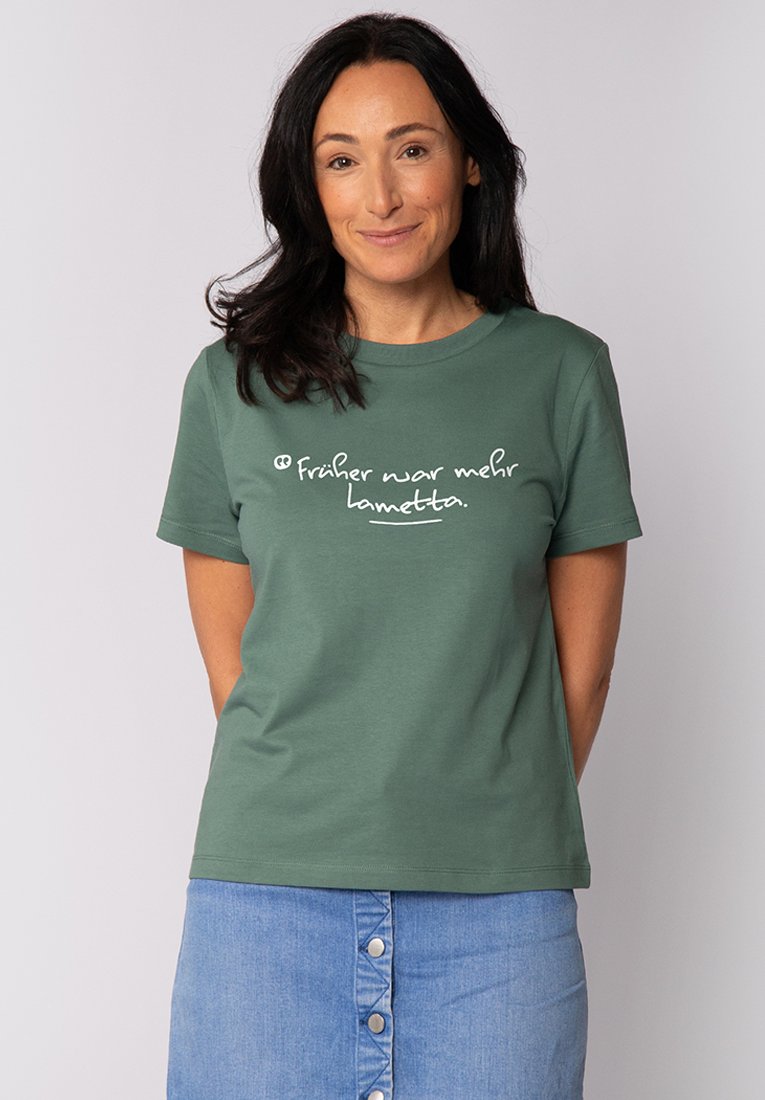Grünes Baumwoll-T-Shirt mit kurzen Ärmeln, das auf der Vorderseite weißem Text mit der Aufschrift "früher war mehr lametta" zeigt. Schlichtes, lässiges Design.