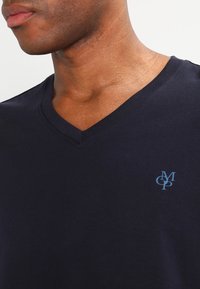 Nærbillede af en person iført en mørkeblå V-hals t-shirt med små lyseblå sammenflettede bogstaver "M," "C" og "P" på brystet.