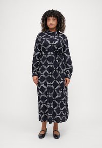 Abito maxi nero con motivo geometrico bianco, maniche lunghe, colletto, apertura frontale con bottoni e vita definita, abbinato a scarpe flat nere.