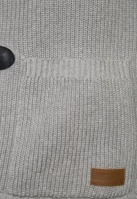 Šedý pletený svetr s velkým černým knoflíkem, žebrovanými manžetami, texturovanou kapsou a malou hnědou koženou �štítkem s raženým textem.