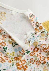 Gros plan sur un vêtement de bébé blanc avec un motif floral et des boutons-pression métalliques le long de l'encolure pour une fixation facile.