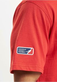 Rød t-skjorte med korte ermer laget av bomull, med et rektangulært merke med teksten "EUROPEAN LEAGUE OF FOOTBALL" i blå og hvite grafikker.