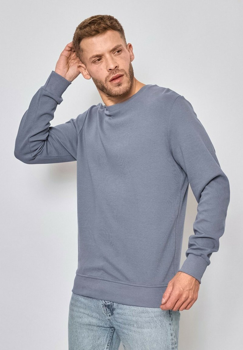 Sweatshirt gris à côtes avec un col rond, des manches longues et des ourlets resserrés, associé à un jean bleu clair. Design simple et décontracté.