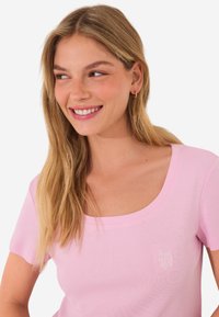 T-shirt rose à manches courtes avec un col en scoop. Présente un petit logo brodé blanc "Saturdays" sur le côté gauche. Texture de tissu lisse.