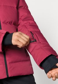 Veste matelassée bordeaux avec un design en piqué, dotée d'une poches à fermeture éclair sur la manche et d'accents noirs aux poignets pour plus de chaleur.