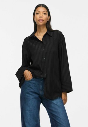 Vrouw draagt een losse zwarte geribde blouse met knopen en brede mouwen en een blauwe spijkerbroek, staand met één hand in haar zak.