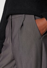 Personne portant un pantalon gris taille haute avec des plis à l'avant et un pull noir texturé, main posée négligemment dans la poche.