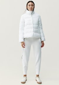 Chaqueta acolchada blanca con cuello alto y diseño acolchado, combinada con pantalones de chándal blancos ajustados con rayas negras en los lados y zapatillas beige.