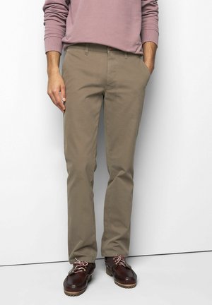 Man wearing beige pants, brown leather shoes with white and red laces, and a mauve long-sleeve shirt, standing against a white wall.