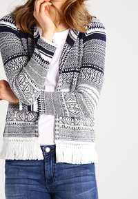 Kvinna i en blå och vit mönstrad stickad cardigan med fransar över vit skjorta och blå jeans, poserar med ena handen nära ansiktet.