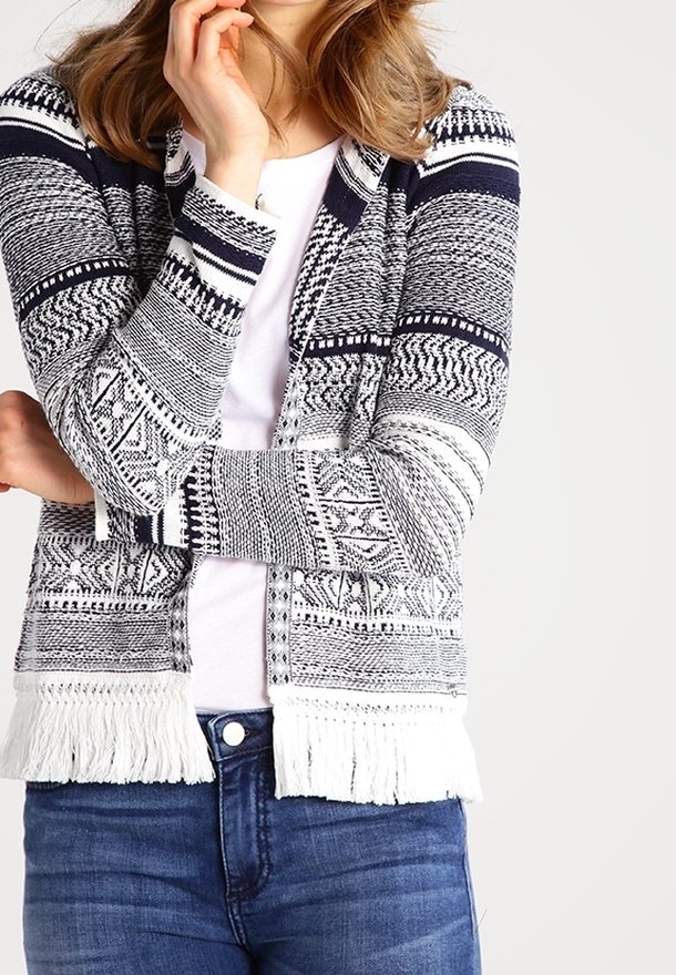 Kvinna i en blå och vit mönstrad stickad cardigan med fransar över vit skjorta och blå jeans, poserar med ena handen nära ansiktet.