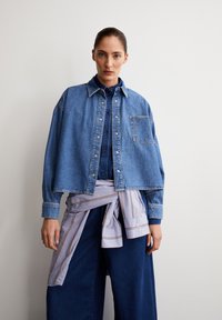 Denim jacket in light blue, featuring a button-down design and a chest pocket. Paired with a patterned shirt tied at the waist and wide-leg jeans.