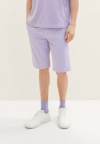 Bermudas de algodón lila con un ajuste relajado y bolsillos laterales, combinadas con zapatillas blancas y calcetines lila a juego. Textura suave, brillo sutil.