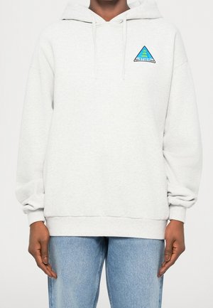 Personne portant un sweat à capuche gris clair avec un petit logo triangulaire bleu et vert sur la partie supérieure gauche de la poitrine et un jean bleu clair.