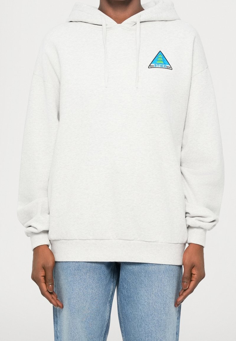 Personne portant un sweat à capuche gris clair avec un petit logo triangulaire bleu et vert sur la partie supérieure gauche de la poitrine et un jean bleu clair.