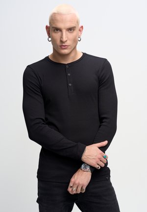 Camicia henley nera a maniche lunghe con tre bottoni, texture a coste, abbinata a pantaloni neri. Gioielli per le mani notevoli includono anelli e un orologio.