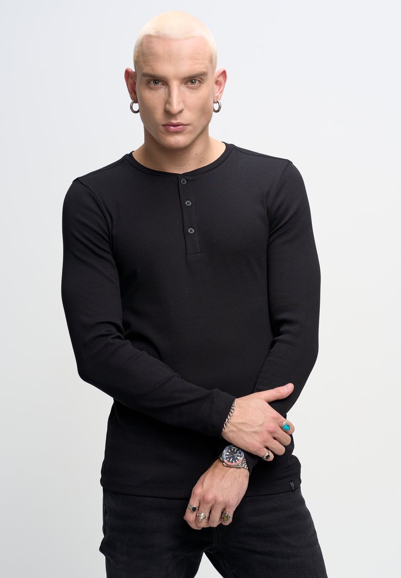 Crna dugorukava henley majica s tri gumba, rebrastog tekstila, u kombinaciji s crnim hlačama. Istaknuti ručni nakit uključuje prstenje i sat.