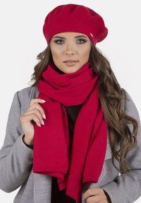 Jeune femme portant un béret rouge et une écharpe rouge assortie, tenant les extrémités de l'écharpe, avec de longs cheveux bruns ondulés, un manteau gris et un maquillage neutre.