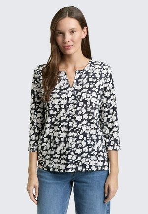 Femme aux longs cheveux bruns portant un chemisier noir et blanc à motif floral avec des manches trois quarts et un jean bleu, debout devant un fond clair uni.