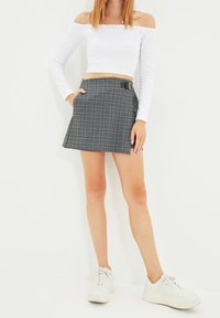 Grauer karierten Minirock mit seitlicher Schnallendetail, kombiniert mit einem weißen schulterfreien Langarmshirt und weißen Turnschuhen. Flache Textur.