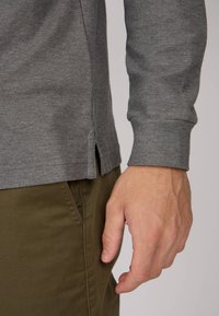 Chemise gris à manches longues avec une finition texturée, fentes latérales à l'ourlet, portée avec un pantalon vert olive, montrant une partie d'une main.