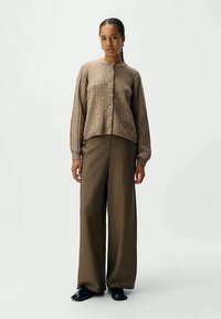 Kvinna som står i en brun kabelstickad knappradad kofta, vida bruna byxor och svarta skor mot en ljus och plain bakgrund.