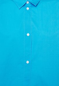 Camicia button-up turchese con una texture liscia, dotata di colletto a punta e bottoni bianchi lungo la patta anteriore.